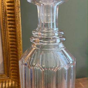 May include: A clear glass decanter featuring a faceted, vertical ribbed body with a starburst pattern at the base. The decanter has a narrow neck with horizontal rings and is topped with a matching, rounded, multi-faceted glass stopper.