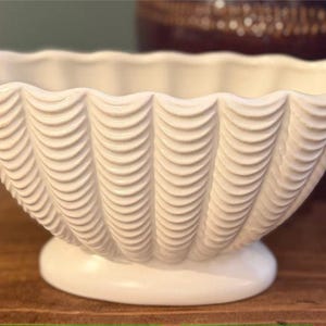 May include: A white ceramic bowl featuring a scalloped, shell-like design with vertical ridges along the exterior. The bowl has an oval-shaped opening with a wavy rim and rests on a smooth, solid oval base, providing a stable foundation for the piece.