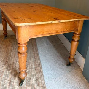 May include: A rectangular wooden table with a warm honey-toned finish, featuring a smooth tabletop with visible wood grain and knots. The table is supported by four thick, turned legs that taper towards the bottom and are fitted with metal castors.