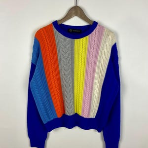 May include: A blue sweater with a colour block design. The sweater features a variety of colours, including blue, orange, grey, yellow, pink, and white. The sweater has a crew neck and long sleeves. The brand name "Versace" is visible on the neckline.