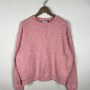 May include: Pink crew neck sweatshirt with a small square patch on the chest. The sweatshirt is hanging on a wooden hanger. The fabric has a heathered texture. The brand name is visible on the neckline.