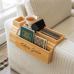 May include: A bamboo sofa arm tray with multiple compartments. It holds a teacup, phone, remote, notebooks, and a small box. The tray is engraved with the name "Olivia Grace". The tray is placed on a striped sofa arm.