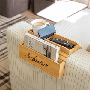Könnte beinhalten: Ein Sofa-Arm-Tablett aus Bambus mit mehreren Fächern. Es enthält eine weiße Tasse mit Kaffee, ein Smartphone, eine Fernbedienung und ein Buch mit dem Titel "Minimalist Home". Das Tablett ist mit dem Namen "Sebastian" graviert.