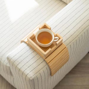 May include: A light-colored wooden sofa arm tray holds a white teacup filled with tea. The tray is designed to fit over the arm of a sofa. The sofa has a white and black striped pattern.