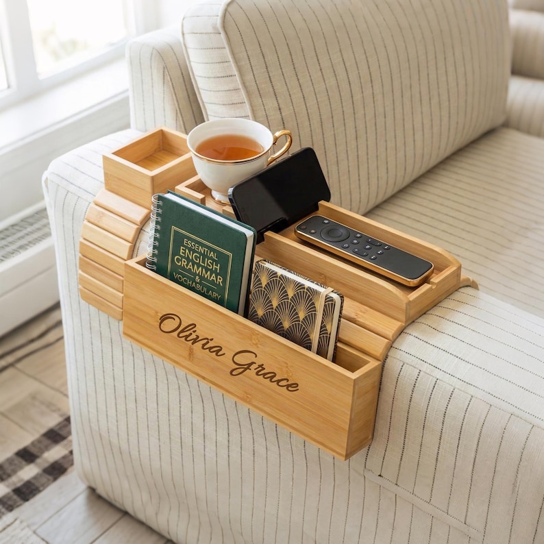 May include: A bamboo sofa arm tray with multiple compartments. It holds a teacup, phone, remote, notebooks, and a small box. The tray is engraved with the name "Olivia Grace". The tray is placed on a striped sofa arm.