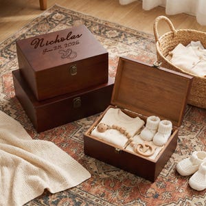 May include: Three dark brown wooden keepsake boxes with metal latches. One box is open, revealing a cream-colored onesie, white knit booties, and a wooden pacifier clip. The top closed box is engraved with the name Nicholas, June 28, 2026, and a pacifier.