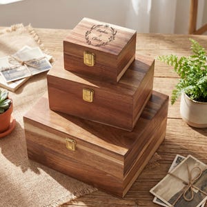 May include: Three wooden storage boxes of different sizes are stacked on a wooden surface. The boxes are a warm brown colour with visible wood grain. The top box has a floral wreath design with the names "Sophia & Claire" engraved on the lid. Each box has a gold-coloured latch.
