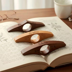 May include: Three wooden book page holders with personalized names: Mia, Seraphina, and Sebastian. Each holder is a different shade of brown and tan, with a floral design and a circular opening. The book is open, displaying text.