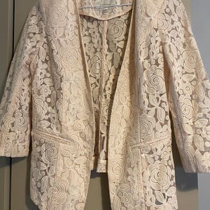 Könnte beinhalten: Ein hellbeige Blazer mit offenem Schnitt und floralem Spitzendesign. Die Jacke hat Dreiviertelärmel und zwei Vordertaschen. Das Spitzendesign zeigt Rosen und andere florale Elemente.
