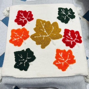 Peut inclure: Un coussin blanc orné de feuilles touffetées de différentes couleurs, dont le rouge, l'orange, le vert et l'or. Le coussin est orné de pompons blancs à chaque coin et est probablement utilisé pour la décoration intérieure.