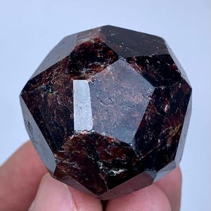 Peut inclure: Un cristal de grenat rouge foncé, facetté et de forme dodécaédrique. La pierre précieuse présente une surface polie et réfléchissante, mettant en valeur sa structure cristalline naturelle. Le cristal est tenu dans une main.