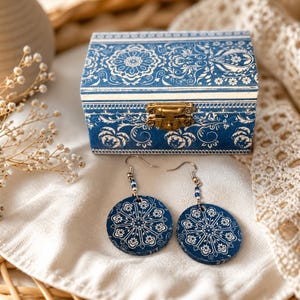 Può includere: Un portagioie blu e orecchini abbinati. La scatola ha un design floreale bianco e una chiusura dorata. Gli orecchini sono rotondi, blu e presentano un motivo floreale bianco. Gli orecchini hanno ganci argentati e dettagli di perline blu.