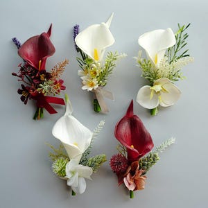 Op de afbeelding: Een assortiment kunstmatige corsages met calla's in wit en bordeaux. Elke corsage bevat groen en accentbloemen, sommige met linten. De achtergrond is effen lichtgrijs.
