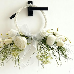 Peut inclure: Une couronne florale blanche avec un anneau perlé, des roses blanches, des tulipes et de la verdure. Un ruban blanc transparent est noué en nœud. La couronne est suspendue à une poignée de porte noire sur un mur blanc.