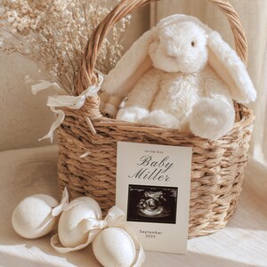 May include: A woven basket holds a plush white bunny and dried flowers. A card with the text "Baby Miller" and an ultrasound image sits in front of the basket, along with three decorative eggs tied with ribbons. The card also reads "September 202X".
