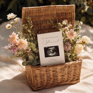 May include: A wicker basket filled with flowers and a card. The card says "We've been keeping a little Secret" and shows an ultrasound image. The text "Arriving January 202x" is also on the card. The flowers are in shades of pink, white, and green.