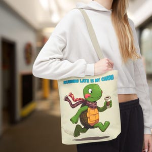Puede incluir: Bolso tote beige con una tortuga de dibujos animados corriendo y sosteniendo una taza de café. La tortuga lleva una bufanda y el texto "RUNNING LATE IS MY CARDIO" es azul. El bolso tiene una correa beige.