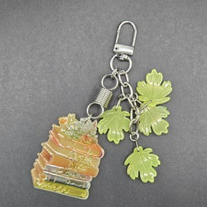May include: A silver-toned metal keychain featuring a stack of books charm with an orange and yellow gradient finish, accented by three light green translucent maple leaf charms attached to a silver chain with a spring-loaded lobster claw clasp.