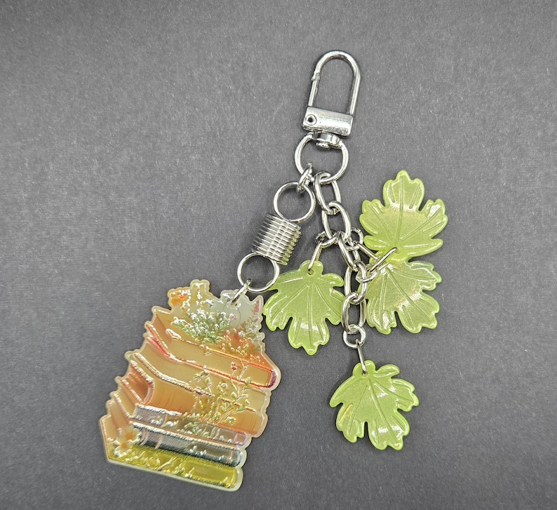 May include: A silver-toned metal keychain featuring a stack of books charm with an orange and yellow gradient finish, accented by three light green translucent maple leaf charms attached to a silver chain with a spring-loaded lobster claw clasp.