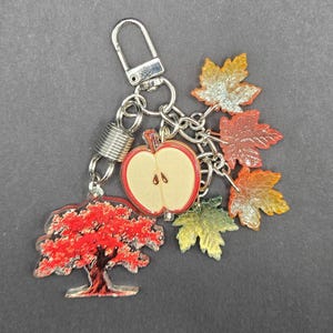 May include: A silver-toned metal keychain featuring a collection of charms including a red tree, a halved apple with a light tan center and red outline, and several translucent maple leaves in shades of orange, yellow, and green attached by rings.