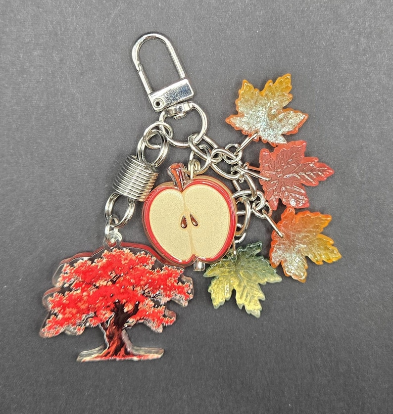 May include: A silver-toned metal keychain featuring a collection of charms including a red tree, a halved apple with a light tan center and red outline, and several translucent maple leaves in shades of orange, yellow, and green attached by rings.
