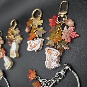 May include: A collection of gold-toned metal keychains featuring charms of autumn-colored maple leaves, small brown acorns, and white rectangular pendants decorated with illustrations of squirrels and mushrooms in shades of orange and brown.