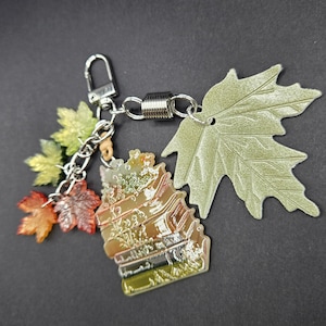 May include: A keychain featuring a stack of books charm with a gradient of green, orange, and brown, accented by a large pale green leaf charm and three smaller translucent maple leaf charms in shades of yellow, orange, and red on a silver chain.