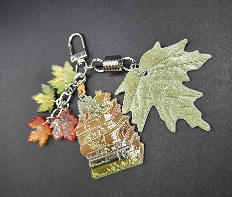 May include: A keychain featuring a stack of books charm with a gradient of green, orange, and brown, accented by a large pale green leaf charm and three smaller translucent maple leaf charms in shades of yellow, orange, and red on a silver chain.