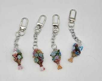 May include: Four keyrings with silver-tone clasps and chains. Each keyring features a bouquet of colourful flowers in a small vase. The flowers are in shades of pink, blue, green, and yellow. The vases are brown and have a pink or yellow ribbon.