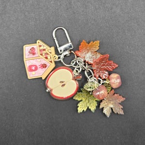May include: A silver-toned metal keychain featuring a pink house charm, a halved apple charm, and several translucent autumn leaf charms in shades of orange, red, and green, along with small round beads attached to a lobster claw clasp.