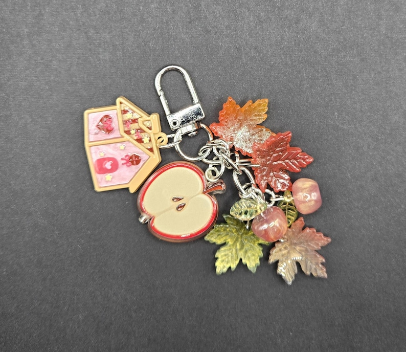 May include: A silver-toned metal keychain featuring a pink house charm, a halved apple charm, and several translucent autumn leaf charms in shades of orange, red, and green, along with small round beads attached to a lobster claw clasp.