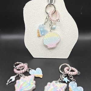 May include: Keychains with a beach theme. Each keychain features a seashell, heart, and starfish charm. The seashells have a pearlescent finish with pastel colors. The keychains are attached to a pink beaded ring.