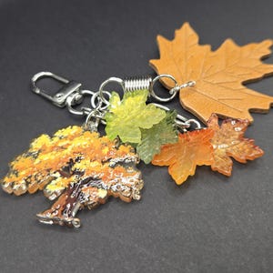 May include: A keychain featuring a silver-toned metal lobster clasp and a spring ring. Attached are several charms: a flat tan leather maple leaf, a translucent green leaf, a translucent orange leaf, and a detailed tree charm with yellow foliage.