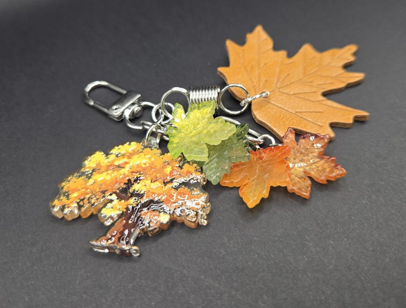 May include: A keychain featuring a silver-toned metal lobster clasp and a spring ring. Attached are several charms: a flat tan leather maple leaf, a translucent green leaf, a translucent orange leaf, and a detailed tree charm with yellow foliage.