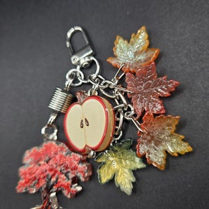 May include: A silver-toned keychain featuring a collection of charms including a red and white apple slice, a red tree, and several translucent maple leaves in shades of orange, yellow, and red, all attached to a metal lobster clasp.