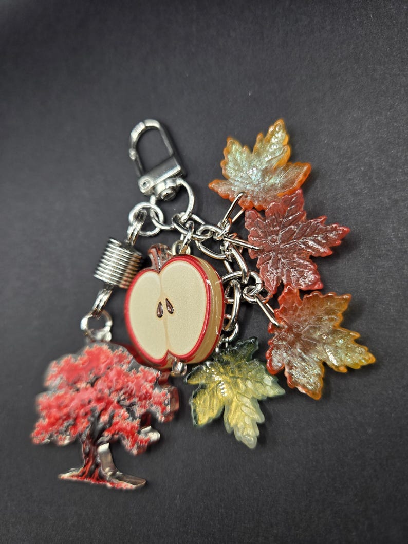 May include: A silver-toned keychain featuring a collection of charms including a red and white apple slice, a red tree, and several translucent maple leaves in shades of orange, yellow, and red, all attached to a metal lobster clasp.