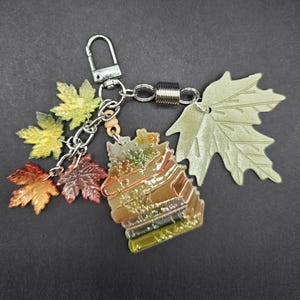 May include: A metal keychain featuring a stack of books charm, three small translucent maple leaf charms in yellow, orange, and brown, and one large matte sage green maple leaf charm, all attached to a silver-toned lobster clasp and chain.