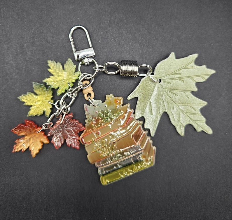 May include: A metal keychain featuring a stack of books charm, three small translucent maple leaf charms in yellow, orange, and brown, and one large matte sage green maple leaf charm, all attached to a silver-toned lobster clasp and chain.