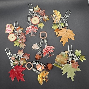 May include: Six keychains featuring autumn-themed charms including maple leaves in red, orange, and green, miniature apples, acorns, trees, stacks of books, and small woodland creatures. Each keychain is attached to a silver-toned metal lobster clasp.