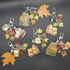 Op de afbeelding: Zes sleutelhangers met herfstthema, waaronder stapels boeken, open boeken, theekopjes en kleine landschapsschilderijen. Elke sleutelhanger is versierd met meerdere esdoornbladbedels van hars in tinten oranje, geel en bruin, bevestigd aan zilverkleurige metalen karabijnsluitingen en spiraalveren.