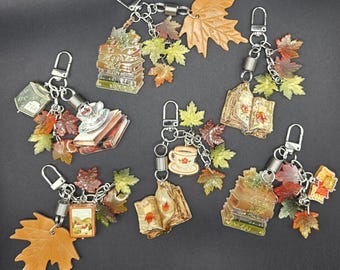 May include: Six keychains featuring autumn-themed charms including stacks of books, open books, teacups, and small landscape paintings. Each keychain is accented with multiple resin maple leaf charms in shades of orange, yellow, and brown, attached to silver-toned metal lobster clasps and coiled springs.