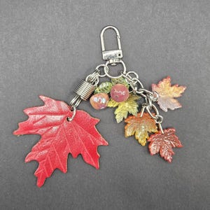May include: A keychain featuring a large red maple leaf charm, several smaller maple leaf charms in shades of orange and yellow, and two small round beads attached to a silver-toned metal lobster clasp and chain link assembly.