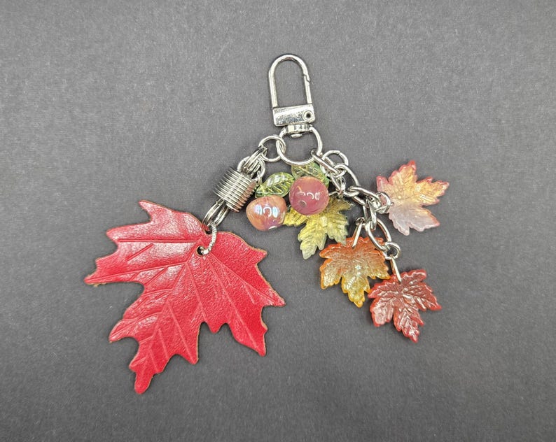May include: A keychain featuring a large red maple leaf charm, several smaller maple leaf charms in shades of orange and yellow, and two small round beads attached to a silver-toned metal lobster clasp and chain link assembly.