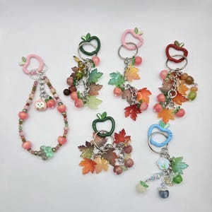 May include: A collection of keychains with apple-shaped clasps in various colors, including pink, green, red, and blue. Each keychain features decorative elements like beads, maple leaves, and small fruit charms. The keychains are arranged on a white surface.