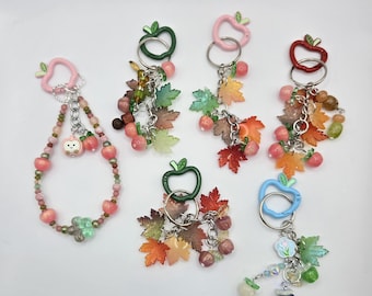 May include: A collection of keychains with apple-shaped clasps in various colors, including pink, green, red, and blue. Each keychain features decorative elements like beads, maple leaves, and small fruit charms. The keychains are arranged on a white surface.