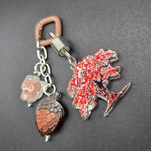 May include: A keychain featuring a light brown plastic carabiner attached to a silver metal spring connector. Three acrylic charms hang from the connector: a small brown squirrel, a textured brown acorn, and a large tree with vibrant red leaves.