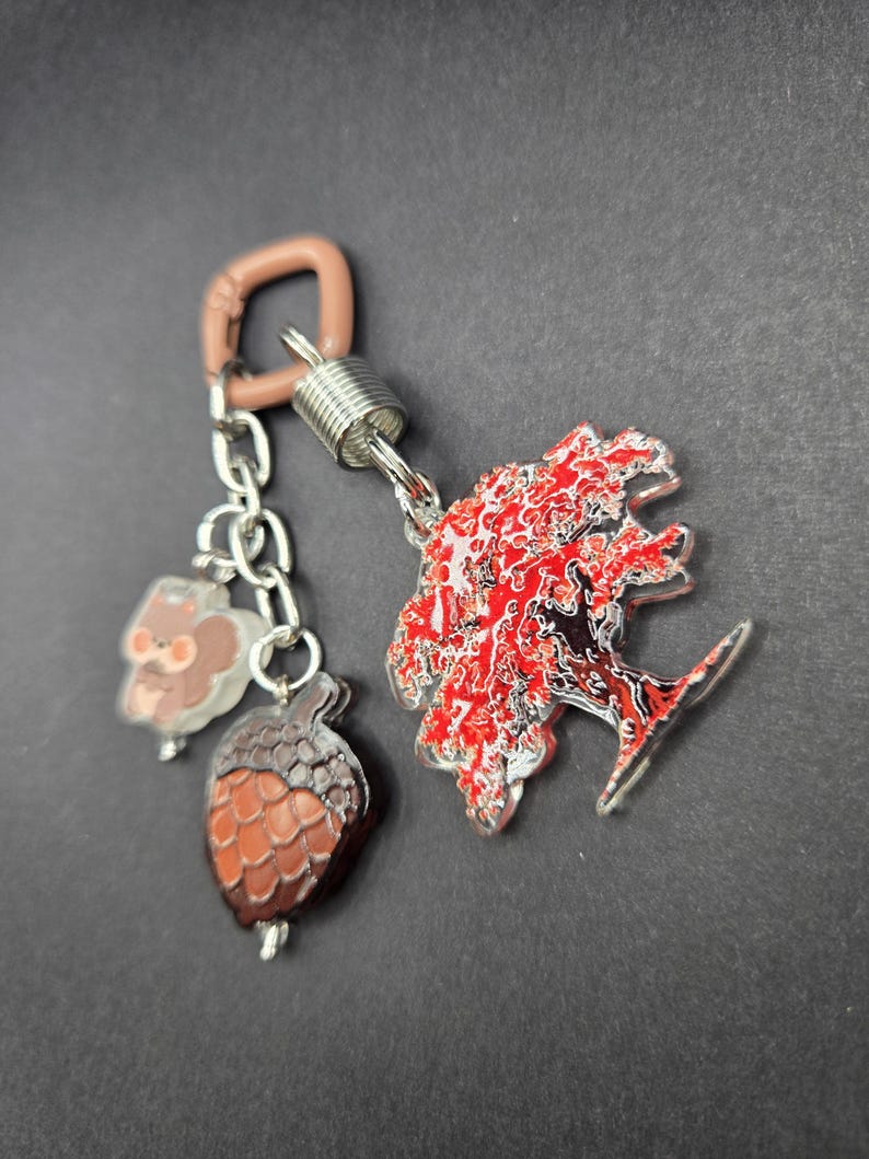 May include: A keychain featuring a light brown plastic carabiner attached to a silver metal spring connector. Three acrylic charms hang from the connector: a small brown squirrel, a textured brown acorn, and a large tree with vibrant red leaves.