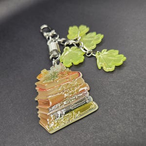 May include: A keychain featuring a stack of books with a metallic finish and a silver-toned metal clasp. Attached to the clasp are three translucent, light green, maple-shaped leaf charms. The book stack displays shades of brown, tan, and gold.
