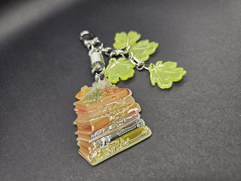 May include: A keychain featuring a stack of books with a metallic finish and a silver-toned metal clasp. Attached to the clasp are three translucent, light green, maple-shaped leaf charms. The book stack displays shades of brown, tan, and gold.