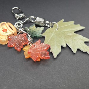 May include: A silver-toned metal keychain featuring a collection of charms including a small beige cup with a red flower design, two translucent red maple leaf charms, one dark green leaf charm, and a larger light green textured maple leaf.